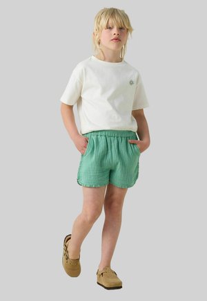 White T-shirt with a small green design, paired with textured mint-green shorts featuring an elastic waistband and side pockets. Brown footwear.
