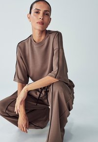 Top marrone oversize con maniche corte e strisce laterali nere, abbinato a pantaloni palazzo coordinati con vita elastica e coulisse.