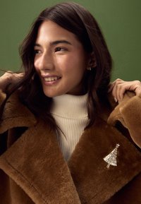 Brown faux fur coat with large lapels, white ribbed turtleneck underneath, and silver Christmas tree brooch on the collar. Green background.