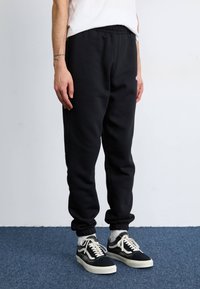 Schwarze Baumwoll-Sweatpants mit elastischem Bund und Bündchen an den Knöcheln. Kombiniert mit schwarz-weißen Turnschuhen, die mit Schnürsenkeln und Logobezügen versehen sind.