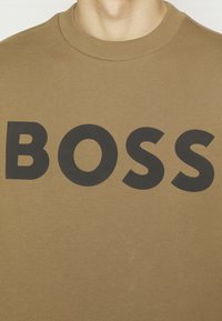 Khaki sweatshirt med rund halsringning och stor svart text "BOSS" över bröstet. Tyget ser mjukt ut med en slät textur.