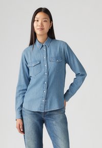 Camisa de denim azul claro com mangas longas, fecho de botões na frente, dois bolsos no peito e colarinho clássico. O tecido tem uma textura suave.