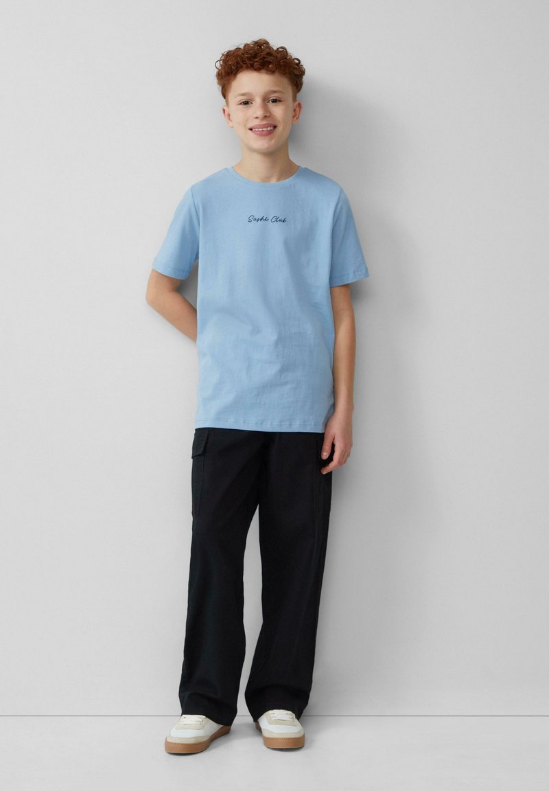 Lichtblauwe katoenen t-shirt met korte mouwen en 'Sophie Clark' geborduurd. Gecombineerd met zwarte cargo-broeken en witte sneakers.