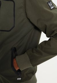 Olivově zelená bunda s ribbovanými manžetami, přední kapsou na zip a náprsní nášivkou s logem a textovými detaily.