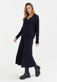 Pull en maille bleu marine avec un col, porté sur une jupe midi assortie. La tenue présente des manches longues et des ourlets côtelés. Des bottes noires épaisses complètent le look.