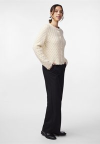 Cremefarbener, gestrickter Cardigan mit einem strukturierten Rautenmuster, rundem Halsausschnitt, fünf Knöpfen und langen Ärmeln, kombiniert mit schwarzen Hosen.