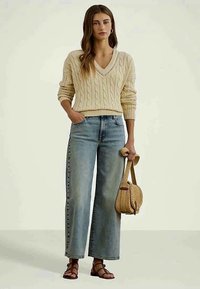 Krēmkrāsas adīts v veida kakla džemperis, gaiši zili platā kāju bikses. Modelis tur austu beige krāsas rokdarbu somu un valkā brūnus sandales ar siksniņām.