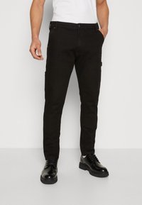 Pantalones negros de corte slim, con bolsillos laterales, costuras en contraste y una textura suave. Combinados con zapatos negros, sobre un fondo neutro.