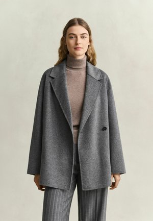 Frau mit langen Haaren, die einen grauen übergroßen Mantel, einen braunen Rollkragenpullover und graue Nadelstreifenhosen vor einem schlichten Hintergrund trägt.