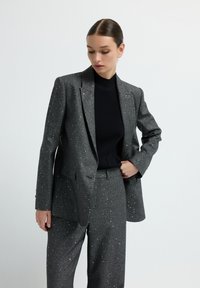 Grauer Blazer und Hosenanzug mit subtilen Glitzer-Muster, ausgestattet mit einem Reverskragen und geradem Schnitt, kombiniert mit einem schwarzen Rippoberteil.