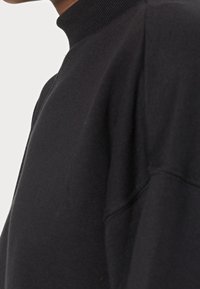 Černá mikina s vysokým rolákem, která se vyznačuje měkkou texturou a volným střihem, zdůrazňující bezešvé provedení a minimální švy.