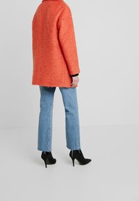 Personne portant un manteau en corail texturé, un jean bleu clair avec des ourlets effilochés et des bottines à talons hauts noires, se tenant devant un fond uni.