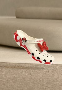 Άσπρο και κόκκινο παπούτσι με πουά και πρόσωπο της Μίνι Μάους με κόκκινο φιόγκο, αιωρούμενο πάνω από μπεζ χαλί σε δωμάτιο με ουδέτερο χρωματικό τόνο.