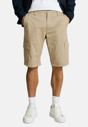 Beige Cargoshorts mit seitlichen Klappentaschen, getragen mit weißen Sneakers, weißen Socken, einem weißen Hemd und einer dunklen Jacke. Hände in den Taschen.