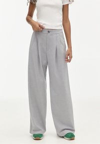 Pantalon gris à jambes larges avec plis, en tissu texturé. Comporte une fermeture à boutons et des poches avant. Porté avec des baskets vertes.