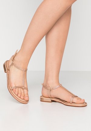 Stopy w różowo-złotych metalicznych sandałach na płaskim obcasie z paskami wokół kostki i palców, stojące na jednolitej białej podłodze na tle białej ściany.