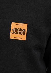 Sudadera negra con un parche de tela naranja en forma de cuadrado que lleva el texto "Jack & Jones" en letras negras en negrita. Textura suave, diseño casual.
