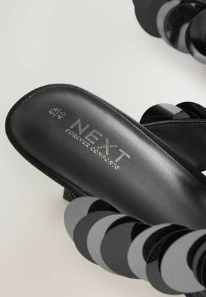 Svarta texturerade sandaler med blanka cirkulära detaljer, med en märkessole som läser "NEXT FOREVER COMFORT" i metallisk skrift.
