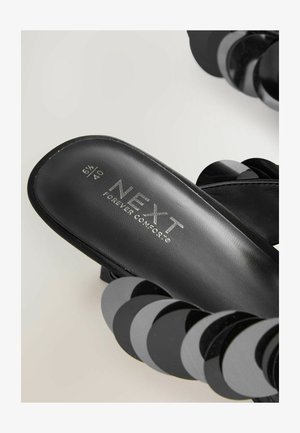 Svarta texturerade sandaler med blanka cirkulära detaljer, med en märkessole som läser "NEXT FOREVER COMFORT" i metallisk skrift.