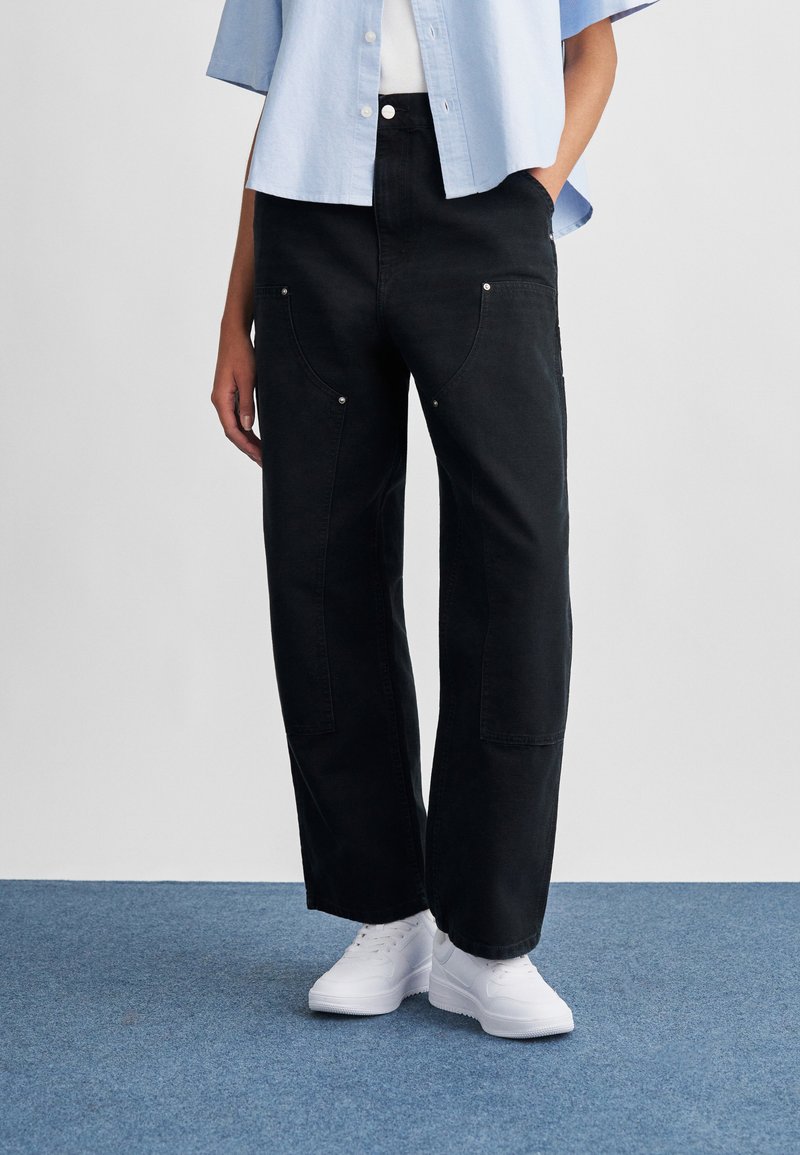 Μαύρο τζιν παντελόνι με φαρδύ πόδι και λεπτομέρειες τσεπών, συνδυασμένο με γαλάζιο πουκάμισο με γιακά και λευκά αθλητικά παπούτσια. Απλός, casual σχεδιασμός.