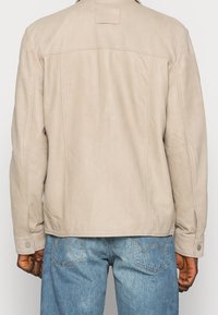 Beige Lederjacke mit glatter Textur, einfachem Design, Knopfmanschetten und einem gestickten Logo-Detail auf dem oberen Rücken.