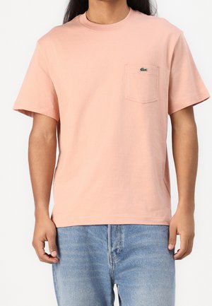 Bladoróżowa bawełniana koszulka z okrągłym dekoltem, krótkimi rękawami, kieszonką na piersi i zielonym logo Lacoste nad kieszonką. Casualowy krój.