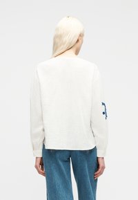 Femme aux longs cheveux blonds portant un chemisier blanc à manches longues avec une broderie bleue sur la manche et un jean bleu, debout de dos à la caméra.