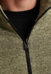 Grøn zip-up sweater med en sort lynlås, lavet af et tekstureret strikstof. Lagt over en sort skjorte. Nærbillede af halsudskæringen.