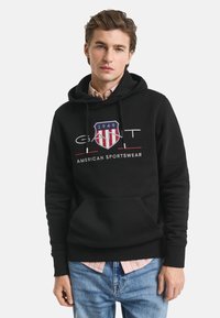 Juodas gobtuvas su priekinėmis kišenėmis, raišteliu ant gobtuvo ir išsiuvinėtu logotipu su raudonu, baltu ir mėlynu skydu. Kasdieniškas kirpimas.