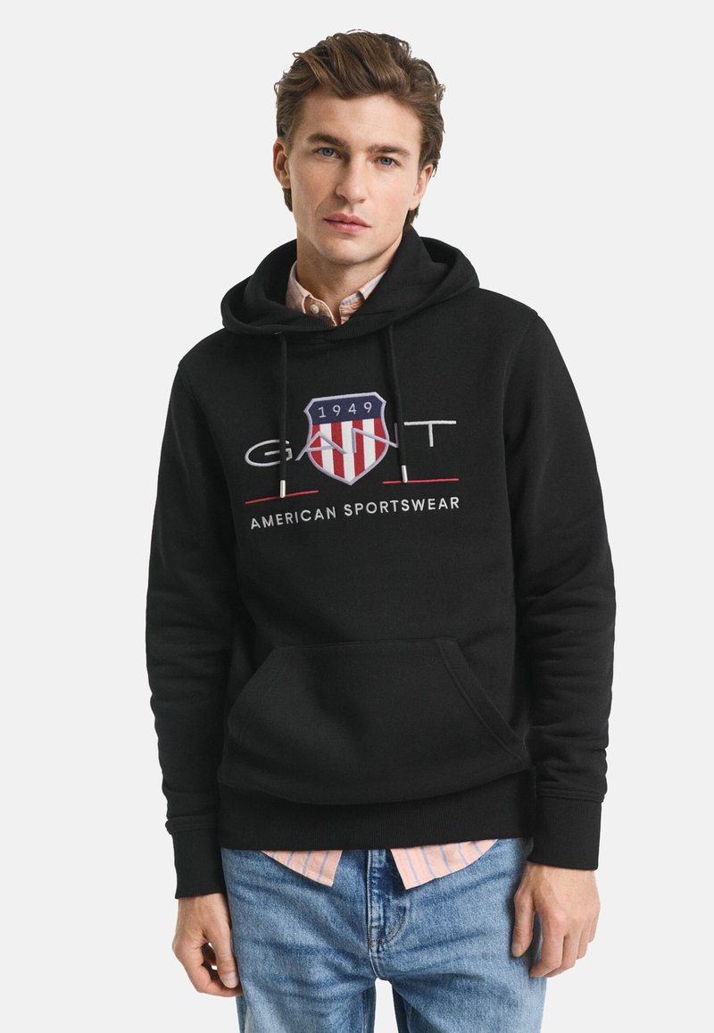 Juodas gobtuvas su priekinėmis kišenėmis, raišteliu ant gobtuvo ir išsiuvinėtu logotipu su raudonu, baltu ir mėlynu skydu. Kasdieniškas kirpimas.