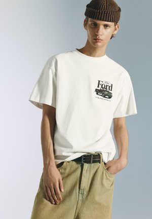 Hvid bomulds t-shirt med et Ford-grafik på venstre bryst, kombineret med lyse beige denimshorts og en brunstribet hue.