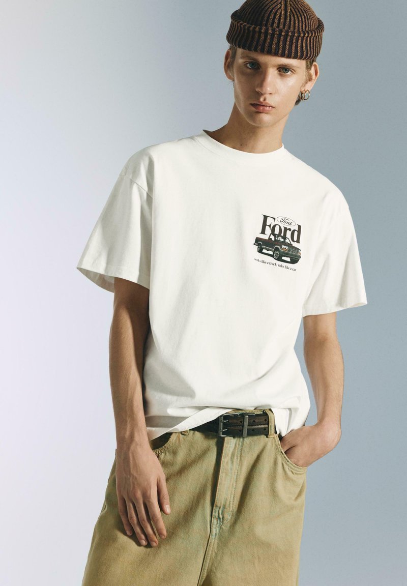 T-shirt en coton blanc avec un motif Ford sur la poitrine gauche, accompagné d'un short en denim beige clair et d'un bonnet rayé marron.