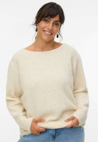 Cream-colored sweater with a textured pattern, wide boat neckline, and long sleeves, paired with denim jeans. Soft and cozy appearance.