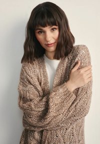 Dzianinowy kardigan w delikatnym odcieniu beżu o grubym splotcie i oversize'owym kroju. Posiada głęboki dekolt w kształcie litery V oraz prążkowane mankiety.