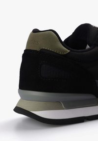 Zapatilla deportiva negra con parte superior de ante y malla, acento verde en el talón y suela de goma en capas en gris, beige y blanco.