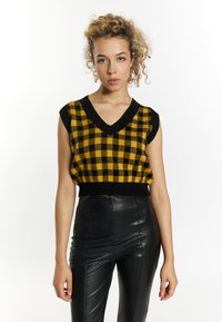 Sárga és fekete kockás kötött crop top V-kivágással és rövid ujjakkal, fekete, magas derekú bőrnadrággal párosítva.