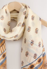 Cream scarf with floral patterns, ribbed texture, and blue-brown accents. Draped gracefully on a wooden hanger.