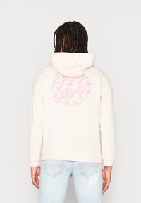 Krämfärgad hoodie med en stor, färgglad grafiktryck på baksidan som har rosa text och gröna accenter. Tillverkad av mjukt tyg.