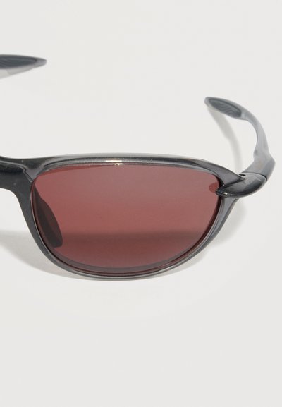 Γυαλιά ηλίου με μαύρο πλαίσιο, καμπυλωτούς βραχίονες και σκουρόχρωμους κόκκινους οβάλ φακούς σε λευκό φόντο.