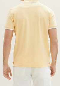 Polo shirt jaune avec bord blanc autour du col et des manches, à manches courtes, fabriqué à partir d'un tissu doux et texturé. Des fentes latérales sont visibles.