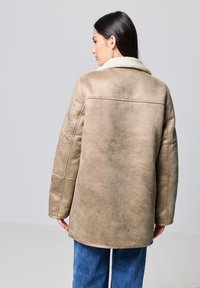 Chaqueta de shearling beige con un cuello de piel, que presenta un corte simple y detalles de costura visibles, llevada sobre pantalones de mezclilla azules.