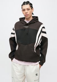 Személy fonott hajjal, fekete-fehér Adidas kapucnis pulóvert és fehér Adidas nadrágot visel, kezét a zsebében tartva egy sima háttér előtt áll.