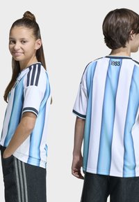 Dva mlada čovjeka u plavo-bijelim prugastim nogometnim dresovima, jedan okrenut prema naprijed s osmijehom, a drugi pokazuje stražnju stranu dresa s natpisom "1893".