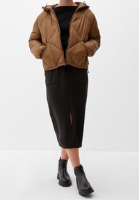 Brązowa pikowana kurtka puchowa z kapturem, zestawiona z czarną sukienką midi z rozcięciem z przodu oraz czarnymi botkami za kostkę.