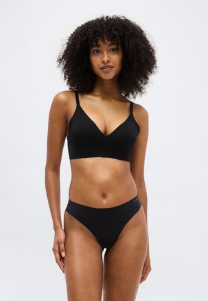 Γυναίκα με σγουρά μαλλιά που φοράει μαύρο ασύρματο σουτιέν και μαύρο εσώρουχο, στέκεται μπροστά σε ένα απλό ανοιχτό φόντο.