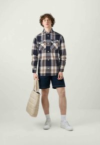 Jovem vestido com uma camisa xadrez azul marinho e bege, calções azul marinho, meias brancas e ténis, segurando uma bolsa de tote acolchoada bege.