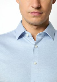 Hellblaues, knopfbares Hemd mit strukturiertem Gewebe, kurzer Kragen und sichtbaren Knöpfen. Nahaufnahme von Kragen und Oberkörper.