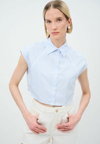 Camicetta a righe blu chiaro, tagliata, con colletto a punta, maniche corte e chiusura a bottoni. Indossata con pantaloncini bianchi a vita alta.