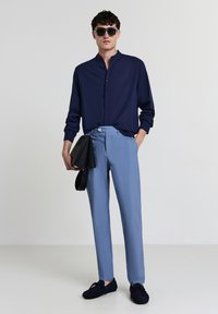Mann in dunkelblauem Hemd mit Mandarin-Kragen, hellblauen Hosen, dunklen Slippern, Sonnenbrille, hält eine schwarze Leder-Clutch. 