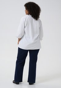 Frau mit lockigem Haar, die eine weiße Bluse mit Streifen und Puffärmeln sowie dunkle Jeans trägt, steht mit dem Rücken zu einem einfarbigen Hintergrund.